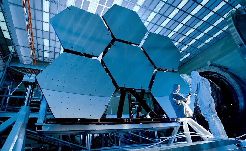 Engineers in protective suits work on telescopic mirrors in a high-tech lab.