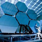 Engineers in protective suits work on telescopic mirrors in a high-tech lab.